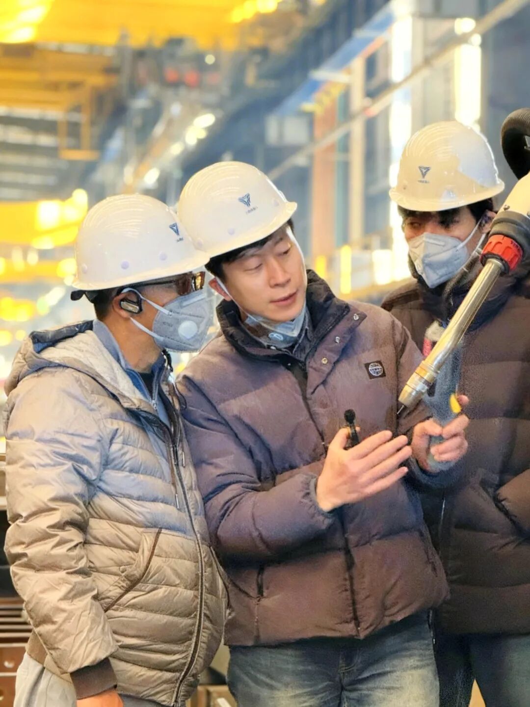 Qiao Zhongliang in a hard hat inspecting a welding tool with workers at a steel plant
